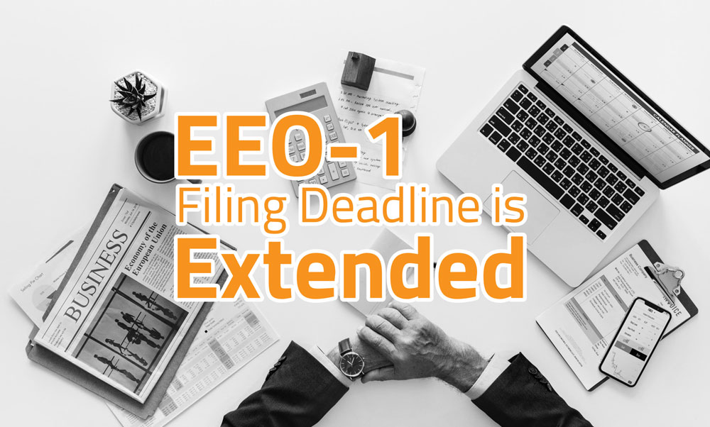 Image of a desk full of business items and a man in a black suit sitting at desk. Words written across the top of image say "EEO-1 Filing Deadline is Extended"